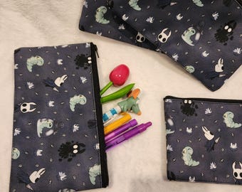 May include: Three gray pencil cases with a black zipper and a white and gray pattern of insects and creatures. The pencil cases are on a white fuzzy surface. The largest pencil case is open and has several colored pencils inside.