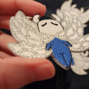 May include: A white embroidered patch with a small, white, winged creature with blue legs. The wings are silver glitter.