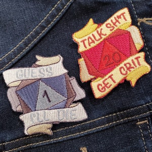 May include: Two embroidered patches on a blue denim background. The first patch is beige with the text "Guess I'll Die" and a purple and gray dice with the number 1. The second patch is yellow with the text "Talk Shit Get Crit" and a red dice with the number 20.