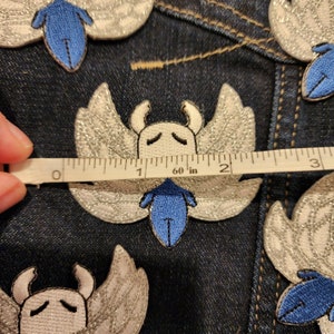 May include: A close-up of a white and silver embroidered patch with a blue insect design. The patch is approximately 1.5 inches wide. The patch is on a blue denim background.
