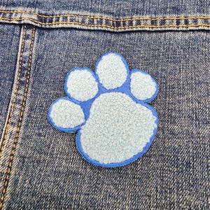 Puede incluir: Un parche de huella de pata azul y blanco con textura peluda. El parche está cosido en una chaqueta vaquera.