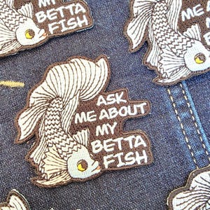 May include: Embroidered patches featuring a stylized betta fish design in white and brown. The patches include the text "ASK ME ABOUT MY BETTA FISH" in a brown font. The patches are on a blue denim background.