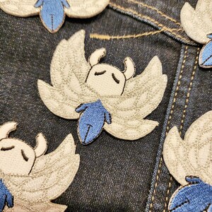 May include: A set of five embroidered iron-on patches featuring a white and blue character with wings. The character has a white face with black eyes and a blue body. The patches are on a blue denim background.