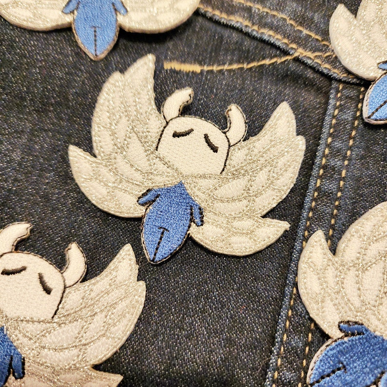 May include: A set of five embroidered iron-on patches featuring a white and blue character with wings. The character has a white face with black eyes and a blue body. The patches are on a blue denim background.