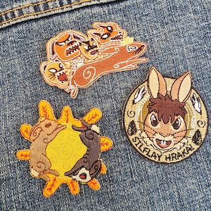 May include: Three embroidered iron-on patches. One features a brown rabbit with the text "Silflay Hraka". Another features a yellow sun with two brown rabbits inside. The last patch features a group of brown and orange cartoon animals.