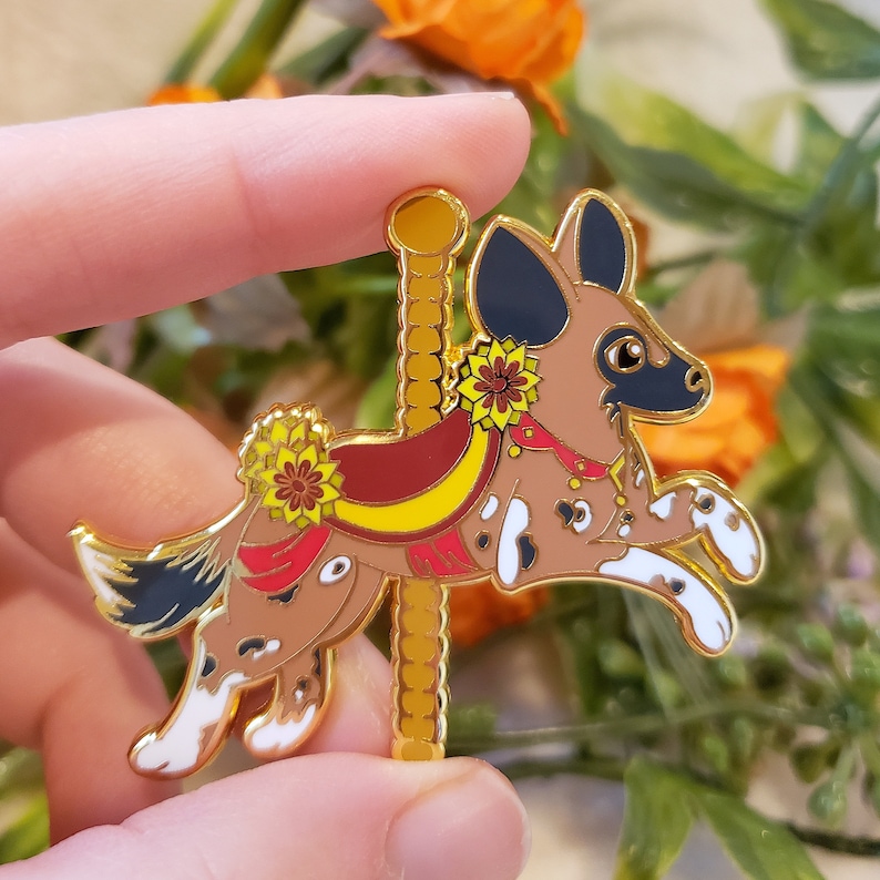 K&ouml;nnte beinhalten: Eine goldfarbene Emaille-Anstecknadel mit einem braunen und schwarzen Afrikanischen Wildhund auf einem Karussellpferd. Der Hund tr&auml;gt einen rot-gelben Sattel mit einem goldfarbenen Blumenakzent.