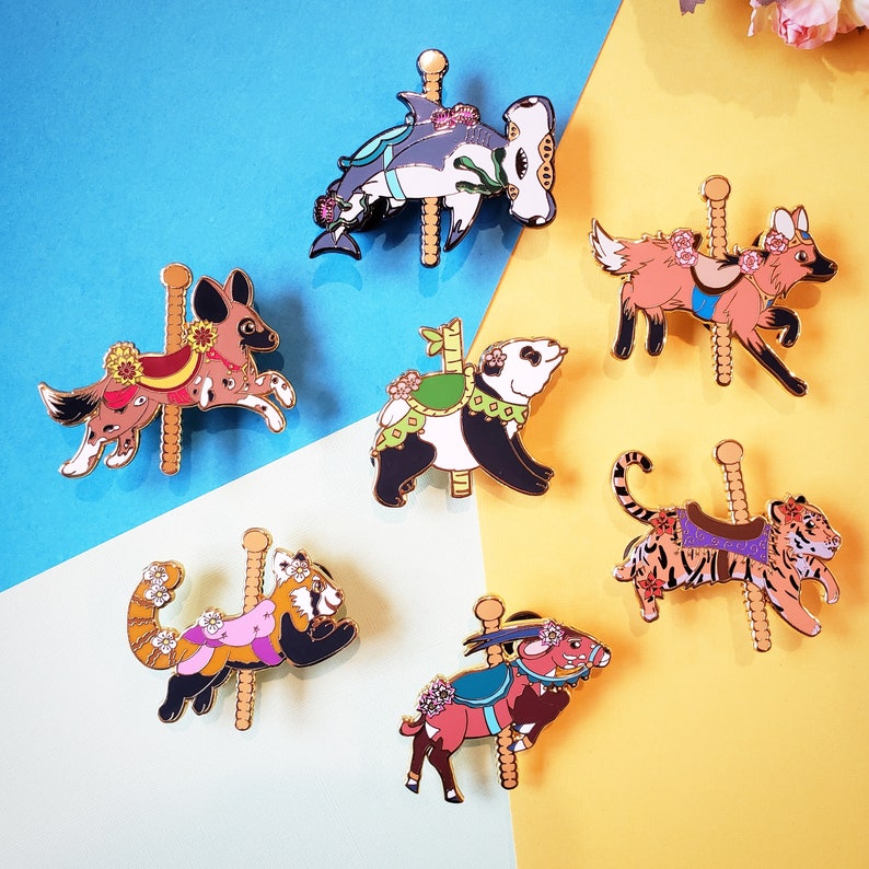 K&ouml;nnte beinhalten: Zehn Emaille-Pins mit verschiedenen Tieren auf einem Karussell. Die Tiere umfassen einen Panda, einen Roten Panda, einen Tiger, einen Hai, einen Hund und einen Fuchs. Die Pins sind alle golden und haben ein farbenfrohes Design.