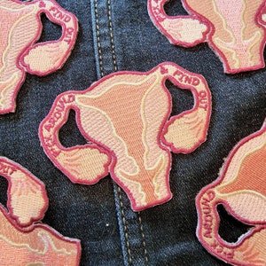 Puede incluir: Cuatro parches bordados que representan una ilustración anatómica estilizada de un útero con el texto "Find Out" y "Get Around" en hilo rosa y blanco sobre un fondo de mezclilla.