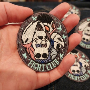 May include: A round, embroidered patch with a dark gray background. The patch features a white character with two black eyes, surrounded by a blue, pink, and white character. The patch has the text "FIGHT CLUB" in a light blue, green, and white font.