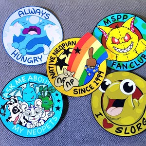May include: A collection of colorful round stickers featuring cartoon characters and text. Designs include a blue creature with "ALWAYS HUNGRY", a yellow monster with "MSPP FAN CLUB", and a green creature with "I LOVE SLORGS".