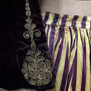 May include: A purple velvet garment with a gold embroidered floral design on the sleeve. The garment is paired with a striped skirt with yellow and purple stripes.