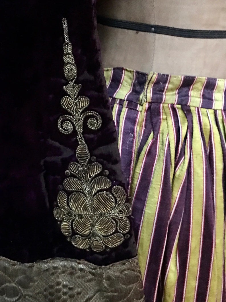 May include: A purple velvet garment with a gold embroidered floral design on the sleeve. The garment is paired with a striped skirt with yellow and purple stripes.