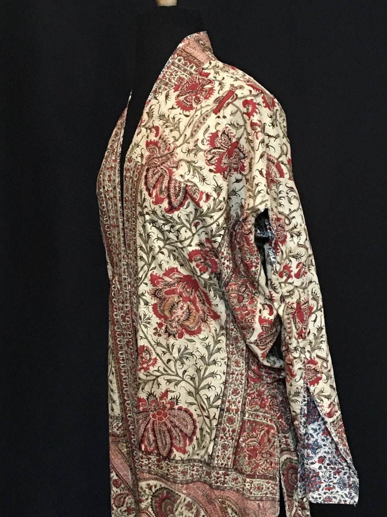 Pu&ograve; includere: Un kimono color crema con stampa floreale rossa e verde. Il kimono ha una silhouette lunga e fluida e un collo largo e aperto.