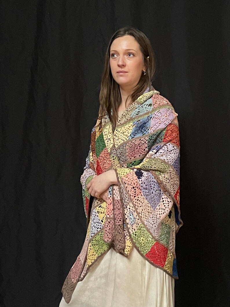 May include: A woman wearing a multi-colored crochet shawl with a granny square pattern. The shawl is made of different shades of pink, green, blue, yellow, and red yarn.