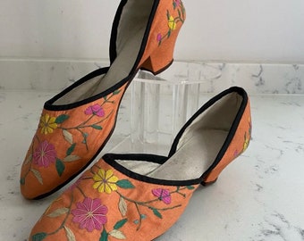 May include: A pair of orange embroidered silk slippers with a black trim and a small heel. The slippers feature a floral design with pink, yellow, and green flowers.