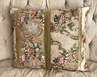 May include: A decorative pillow with a floral pattern in shades of pink, red, green, and blue. The pillow has a beige background with gold and green accents. The pillow is sitting on a white tufted sofa.