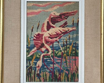 May include: A framed needlepoint embroidery depicting two pink flamingos in flight over a blue body of water with green reeds. The background is a light blue sky with pink clouds.