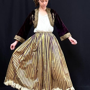 May include: A woman wearing a long, yellow and burgundy striped skirt with a white lace trim. She is also wearing a burgundy velvet jacket with gold embroidery and a white blouse.