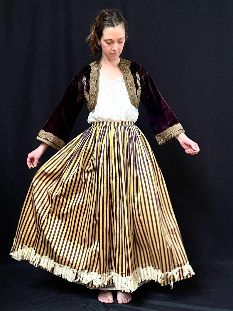 May include: A woman wearing a long, yellow and burgundy striped skirt with a white lace trim. She is also wearing a burgundy velvet jacket with gold embroidery and a white blouse.