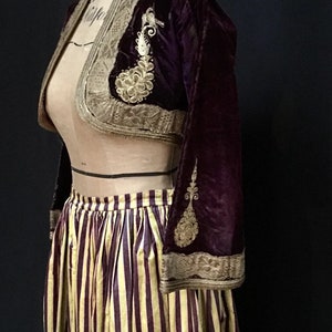 May include: A burgundy velvet cropped jacket with gold embroidery and a matching trim. The jacket is paired with a striped skirt with yellow and burgundy stripes.