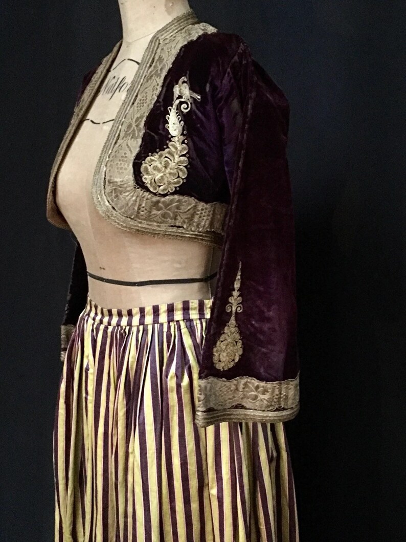 May include: A burgundy velvet cropped jacket with gold embroidery and a matching trim. The jacket is paired with a striped skirt with yellow and burgundy stripes.