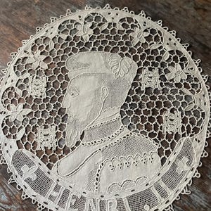 May include: Cream-colored, circular lace doily featuring a profile portrait of a man with a beard and the name "HENRI" in a banner. The intricate design includes floral and geometric patterns, with a scalloped edge.