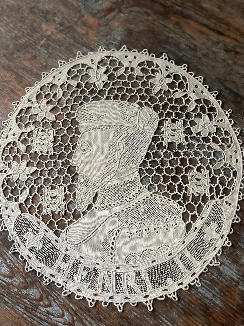 May include: Cream-colored, circular lace doily featuring a profile portrait of a man with a beard and the name "HENRI" in a banner. The intricate design includes floral and geometric patterns, with a scalloped edge.