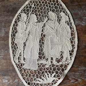 May include: Oval-shaped, off-white lace doily featuring a detailed depiction of several figures in period clothing. The lace has an open, honeycomb-like pattern around the edges, and the figures are intricately rendered.