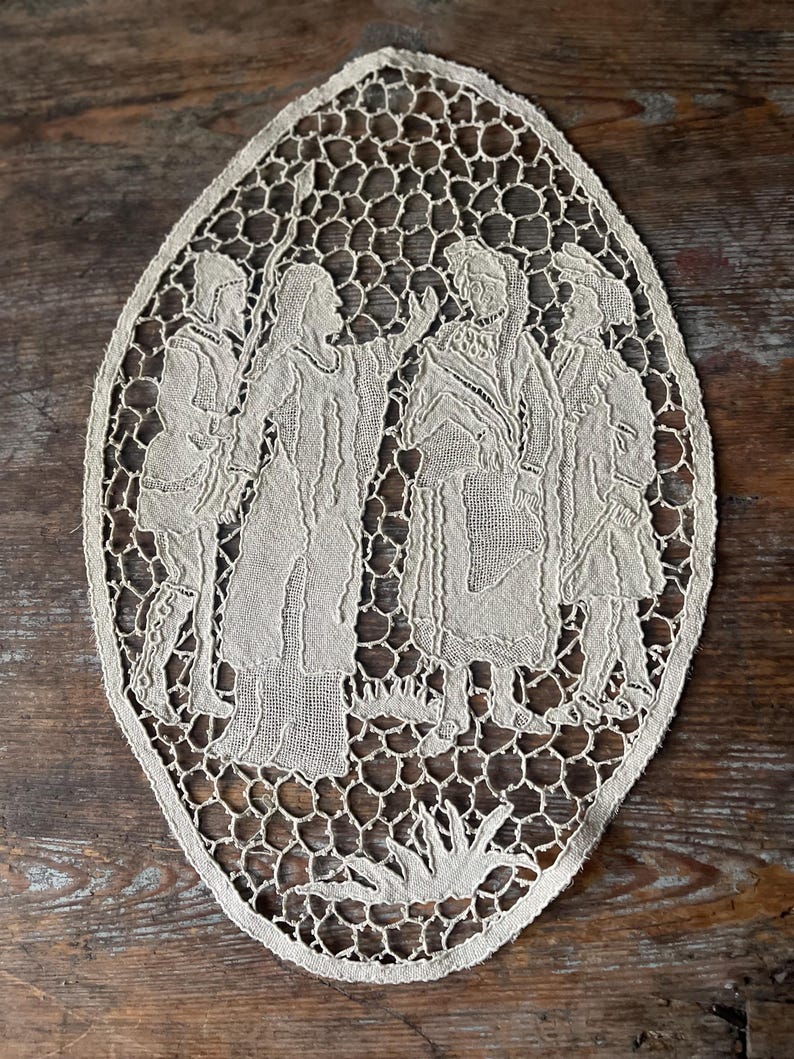 May include: Oval-shaped, off-white lace doily featuring a detailed depiction of several figures in period clothing. The lace has an open, honeycomb-like pattern around the edges, and the figures are intricately rendered.