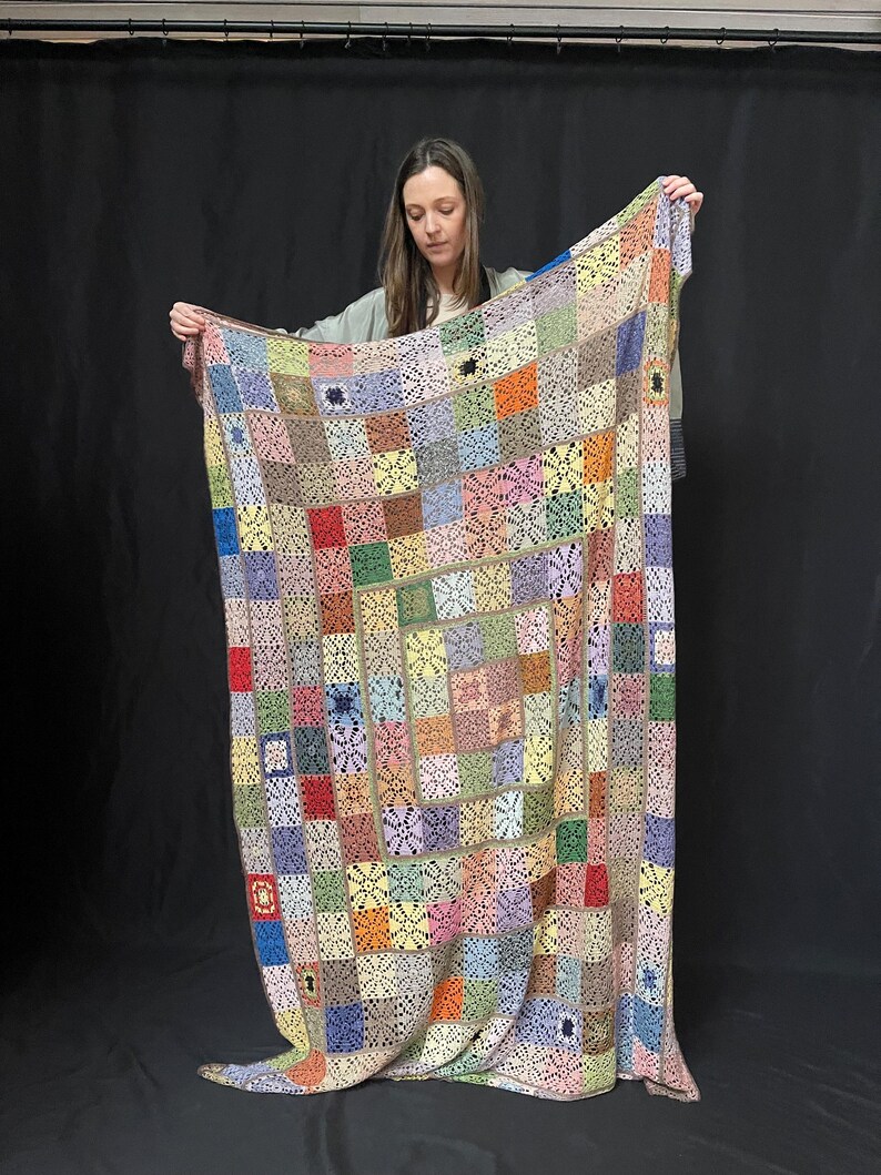 May include: A large, colorful crocheted blanket with a patchwork design. The blanket is made up of squares in various colors, including red, orange, yellow, green, blue, purple, and brown. The squares are joined together with a white crocheted border.