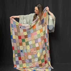 May include: A large, colorful crochet blanket with a patchwork design. The blanket is made up of squares in various colors, including red, green, blue, yellow, and pink. The blanket is being held up by a person, who is wearing a long-sleeved shirt and pants.