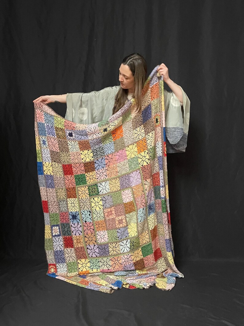 May include: A large, colorful crochet blanket with a patchwork design. The blanket is made up of squares in various colors, including red, green, blue, yellow, and pink. The blanket is being held up by a person, who is wearing a long-sleeved shirt and pants.