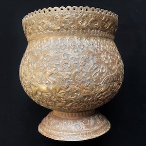 Może przedstawiać: Złota metalowa doniczka z misternymi wzorami kwiatowymi i zwierzęcymi. Doniczka ma szeroką, okrągłą podstawę i falisty brzeg.