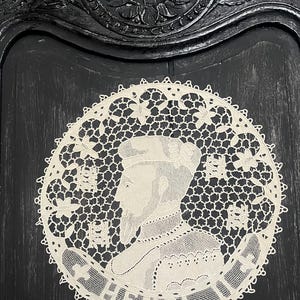 May include: A cream-colored, circular lace doily featuring a profile portrait of a man with the name "HENRY" at the bottom. The doily has a delicate, openwork design with floral and geometric patterns. The background is a dark, textured surface.