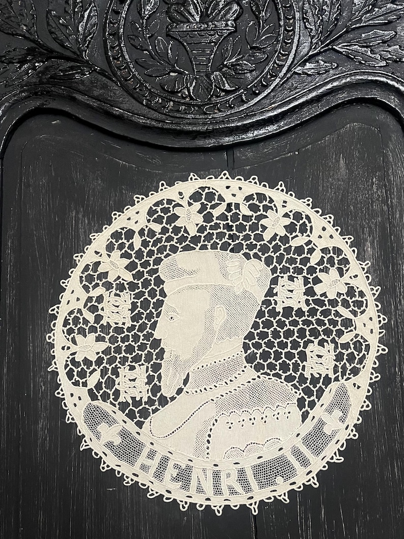 May include: A cream-colored, circular lace doily featuring a profile portrait of a man with the name "HENRY" at the bottom. The doily has a delicate, openwork design with floral and geometric patterns. The background is a dark, textured surface.