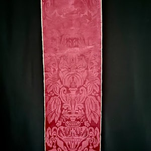 May include: A close-up of a deep red damask fabric with a white border. The fabric features a repeating pattern of a large, ornate vase with floral motifs.