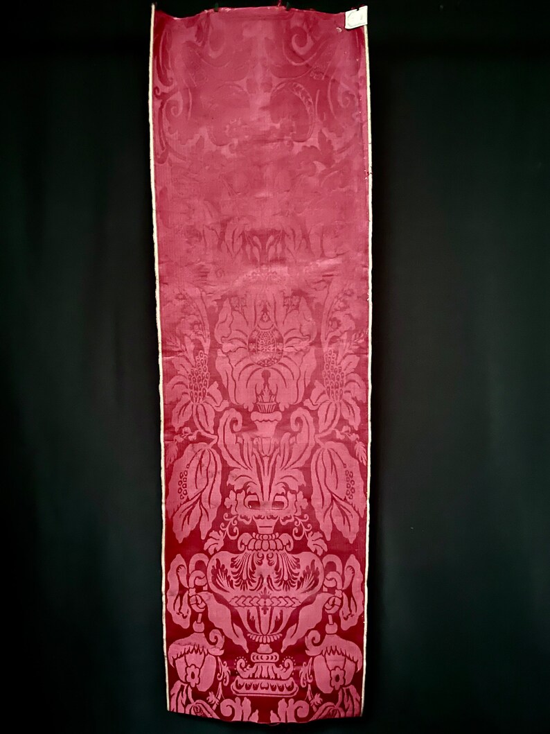 May include: A close-up of a deep red damask fabric with a white border. The fabric features a repeating pattern of a large, ornate vase with floral motifs.