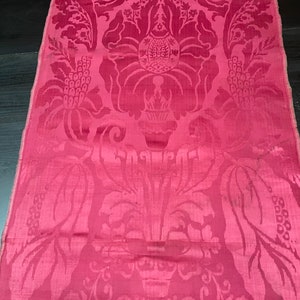 May include: A close-up of a pink damask fabric with a floral pattern. The fabric has a subtle sheen and a luxurious feel.