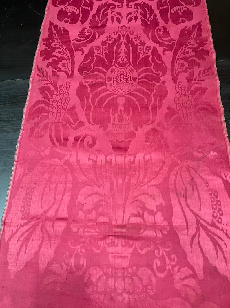 May include: A close-up of a pink damask fabric with a floral pattern. The fabric has a subtle sheen and a luxurious feel.