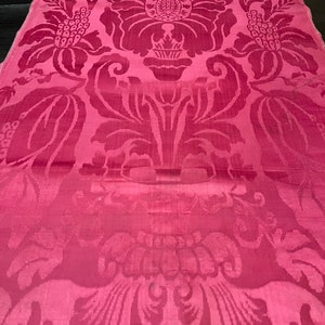 May include: A close-up of a pink fabric with a damask pattern. The pattern features intricate floral designs and swirls.