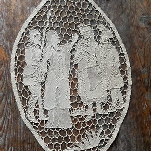 May include: Oval-shaped, off-white lace doily featuring a detailed scene of several figures in period clothing. The lace has an open, honeycomb-like pattern around the figures. The figures appear to be engaged in a conversation.