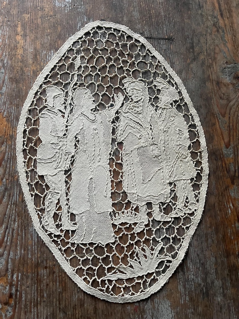 May include: Oval-shaped, off-white lace doily featuring a detailed scene of several figures in period clothing. The lace has an open, honeycomb-like pattern around the figures. The figures appear to be engaged in a conversation.