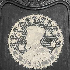 May include: A cream-colored, circular lace doily featuring a profile portrait of a man with the name "HENRI" in a banner. The doily has a detailed floral and geometric openwork design, set against a dark, textured background.