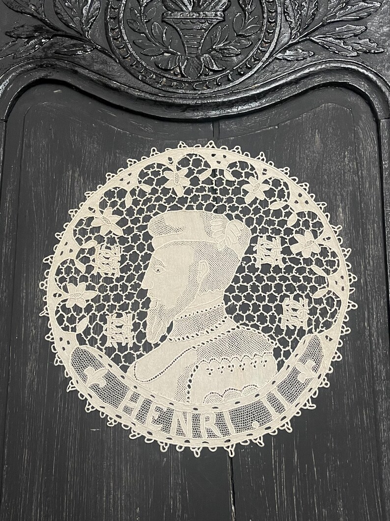 May include: A cream-colored, circular lace doily featuring a profile portrait of a man with the name "HENRI" in a banner. The doily has a detailed floral and geometric openwork design, set against a dark, textured background.