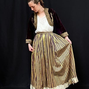May include: A woman wearing a long, striped skirt with a gold and burgundy pattern. The skirt has a lace trim and is paired with a velvet jacket with gold embroidery.
