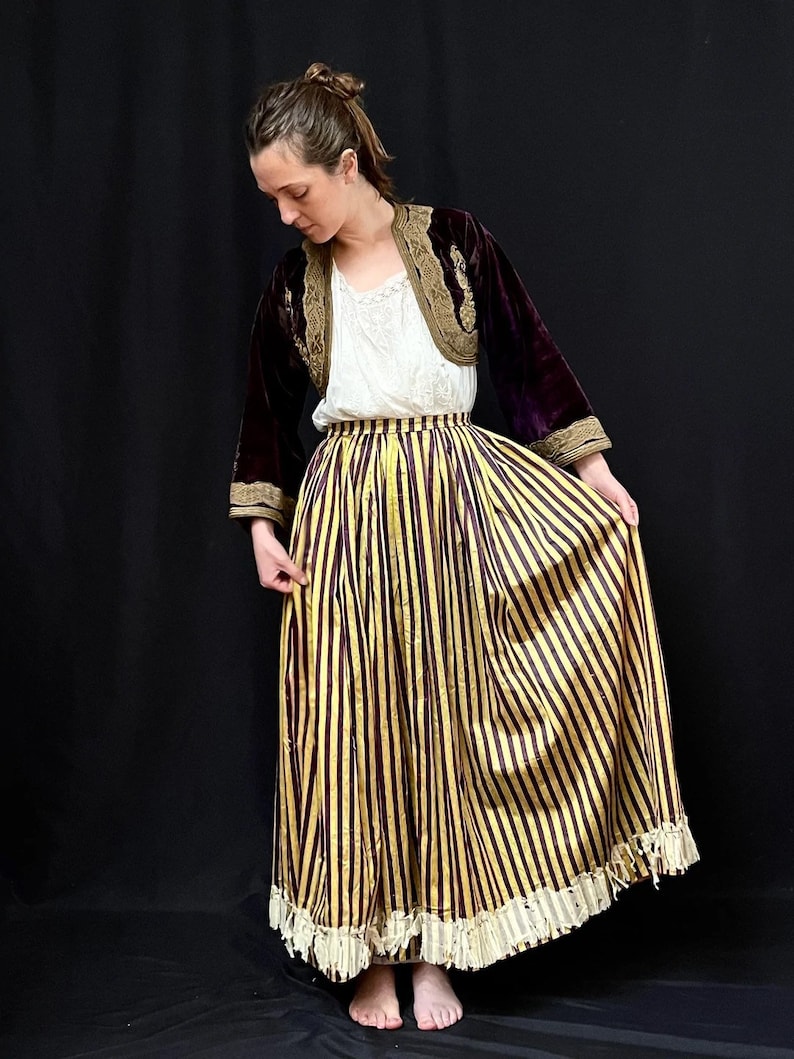 May include: A woman wearing a long, striped skirt with a gold and burgundy pattern. The skirt has a lace trim and is paired with a velvet jacket with gold embroidery.