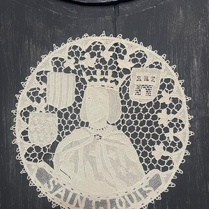 May include: A cream-colored, circular lace doily featuring a portrait of a crowned figure, possibly a king, with the text "SAINT LOUIS" in a banner below. The design includes heraldic shields and intricate lace patterns.