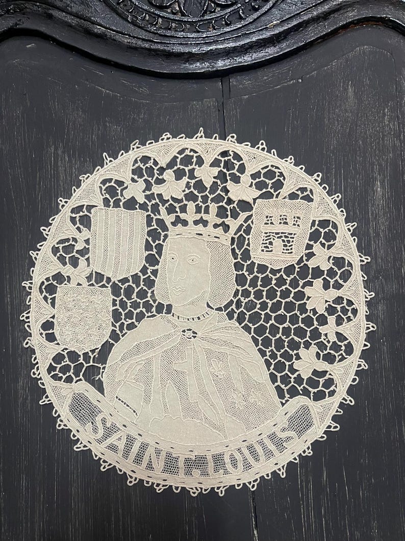 May include: A cream-colored, circular lace doily featuring a portrait of a crowned figure, possibly a king, with the text "SAINT LOUIS" in a banner below. The design includes heraldic shields and intricate lace patterns.