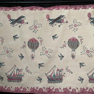 Könnte beinhalten: Cremefarbener Stoff mit einem sich wiederholenden Muster aus Flugzeugen, Heißluftballons und Schiffen in Grau-, Türkis- und Rosatönen. Das Design umfasst florale Akzente und fliegende Vögel mit einem dekorativen Rand.
