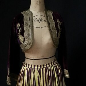 May include: A purple velvet jacket with gold embroidery and lace trim, paired with a yellow and purple striped skirt. The jacket has long sleeves and a cropped length. The skirt is full and has a high waist.